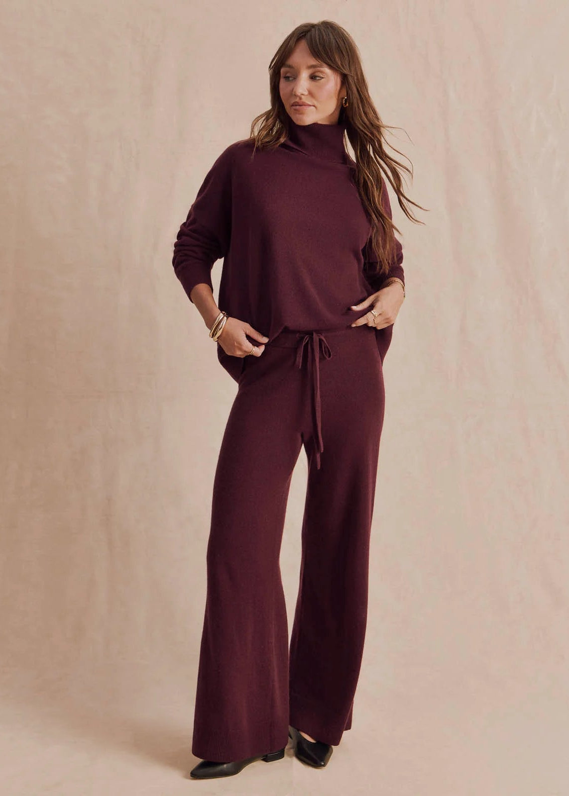 A woman modeling a matching burgundy knit two-piece set consisting of a long-sleeve mock-neck top and wide-leg pants with a drawstring waist, set against a textured beige studio backdrop.