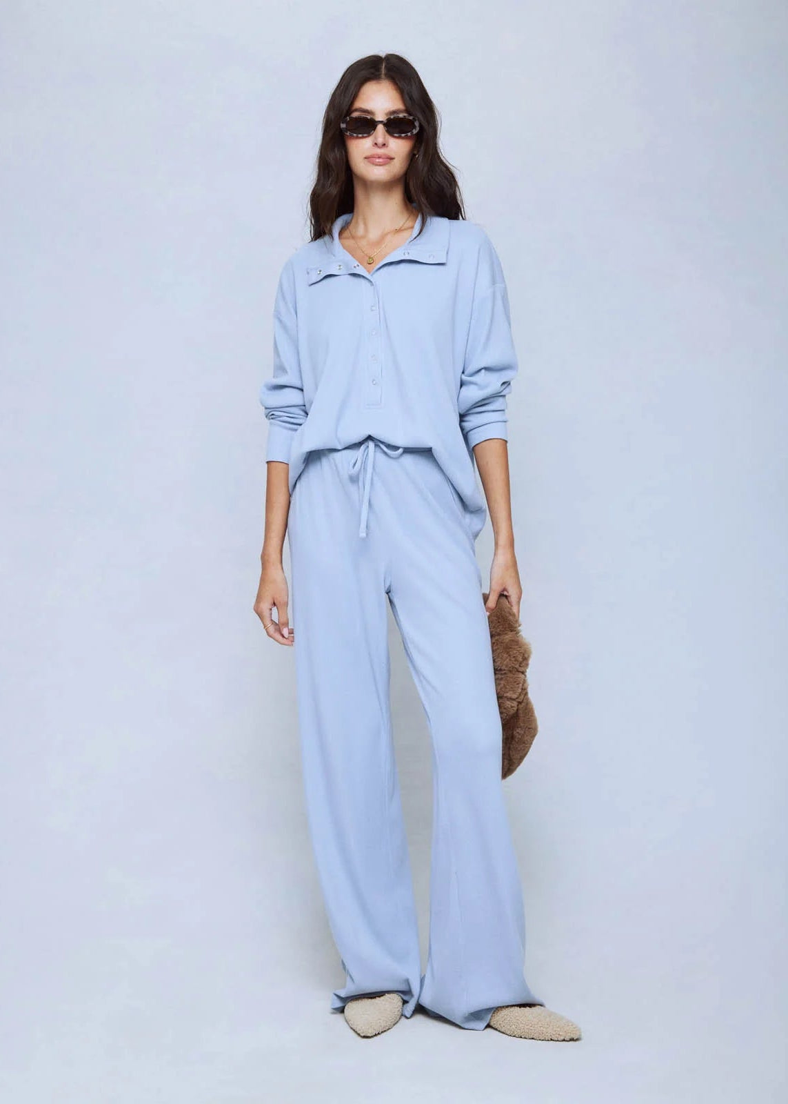 A woman modeling a light blue two-piece lounge set consisting of a long-sleeve collared top and matching wide-leg pants, styled with sunglasses, a brown textured bag, and beige slippers against a light gray studio background.