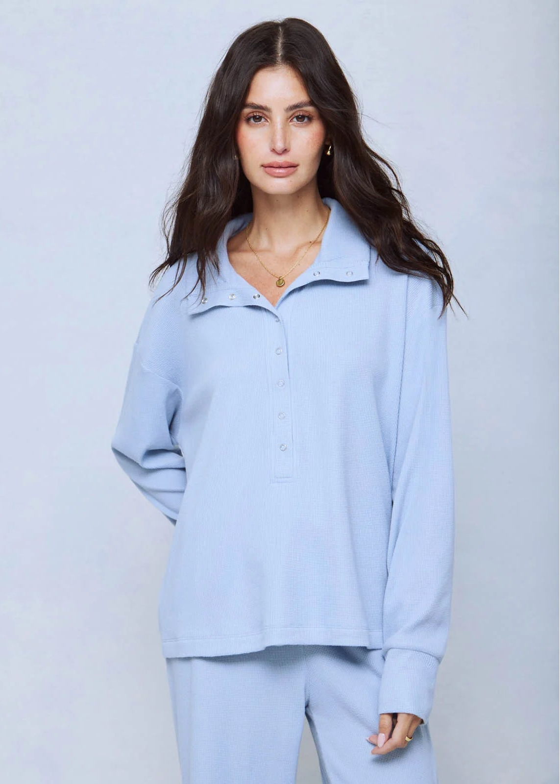 A woman modeling a light blue long-sleeve collared top with a short button placket, paired with matching light blue pants, against a light gray studio background.