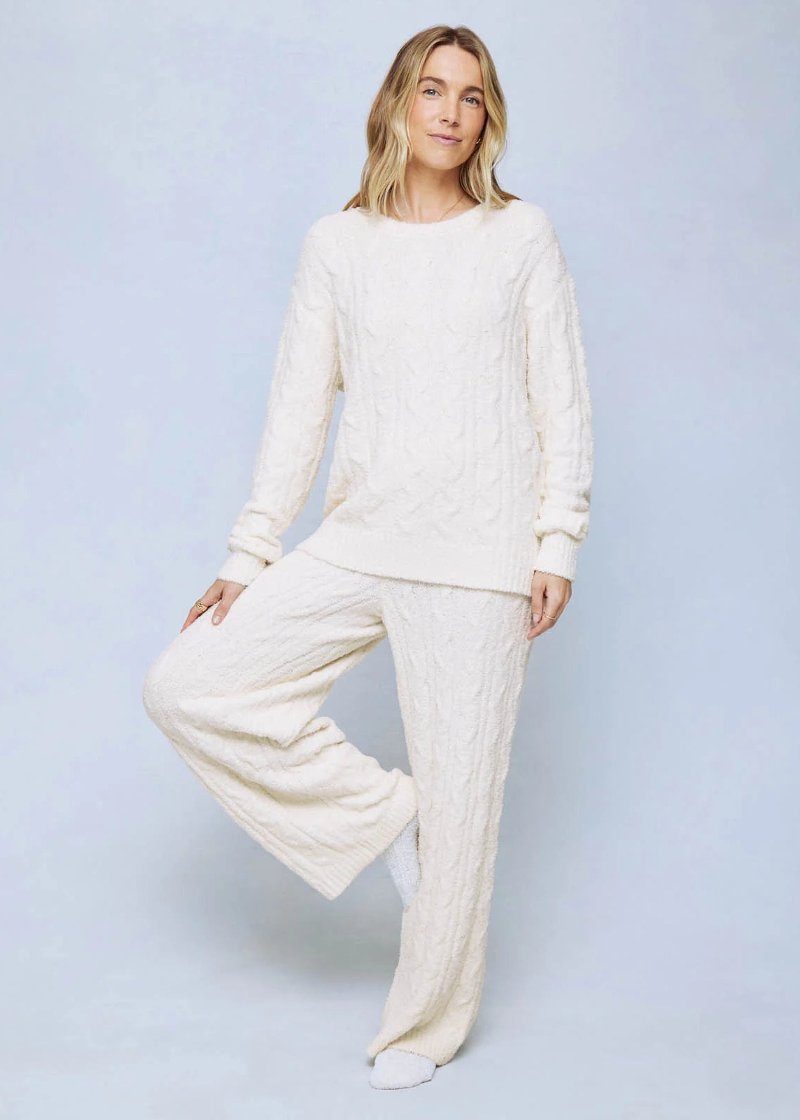 A woman modeling a matching cream cable-knit lounge set, consisting of a long-sleeve top and wide-leg pants, against a light blue studio background.