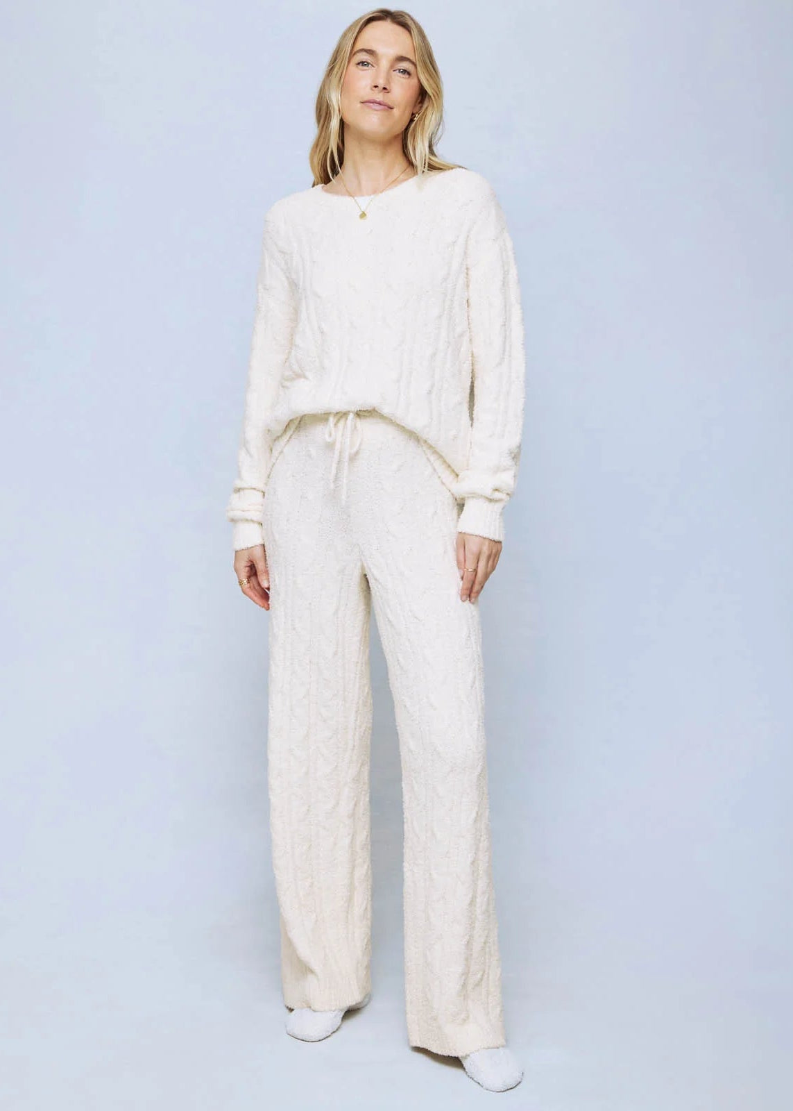 A woman modeling a matching cream-colored cable-knit lounge set, consisting of a long-sleeve top and wide-leg pants, against a light blue studio background.