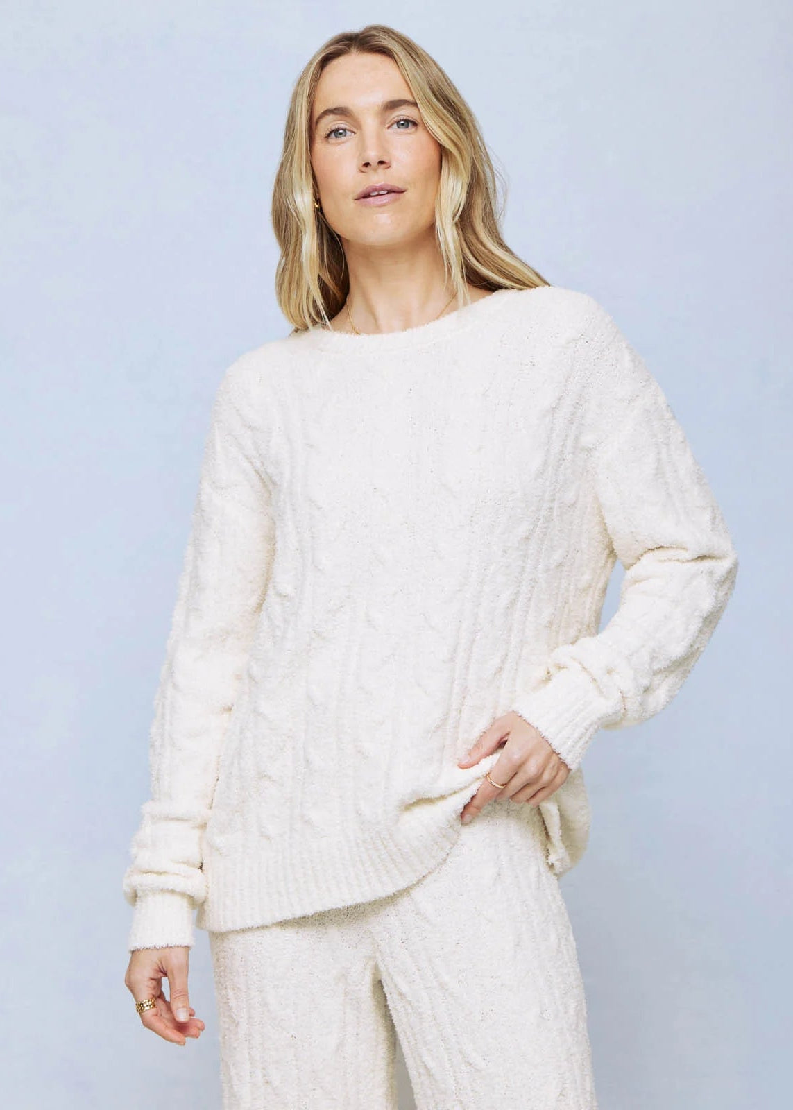 A woman modeling a cream cable-knit crewneck sweater and matching knit pants against a light blue studio background.