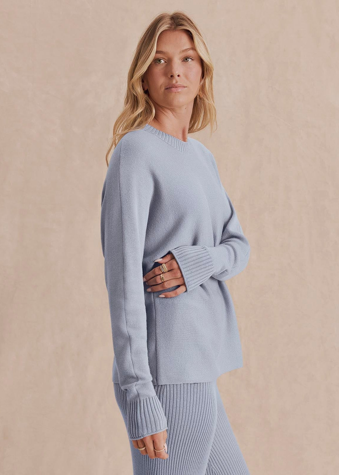 A woman wearing a light blue ribbed knit sweater and matching ribbed knit pants, standing against a warm beige textured background.