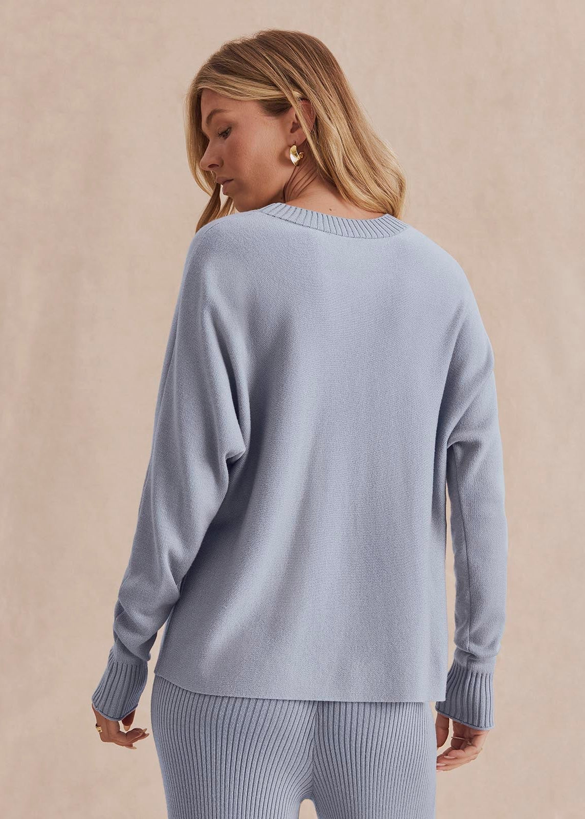 A woman shown from the back wearing a light blue knit sweater and matching ribbed pants against a warm beige studio backdrop.