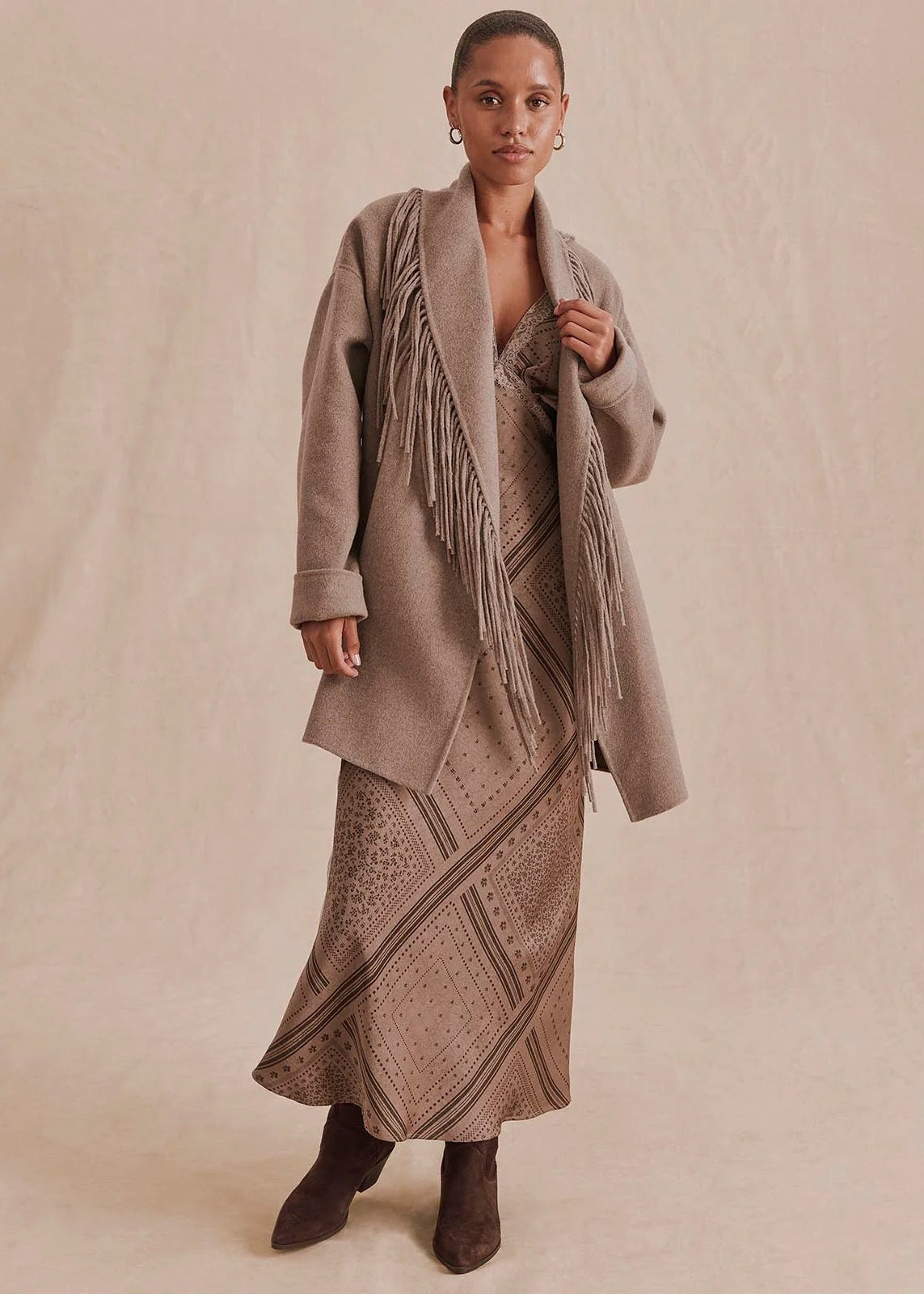 A model stands against a warm beige backdrop wearing a taupe open-front fringed coat over a matching patterned maxi dress and dark brown ankle boots.