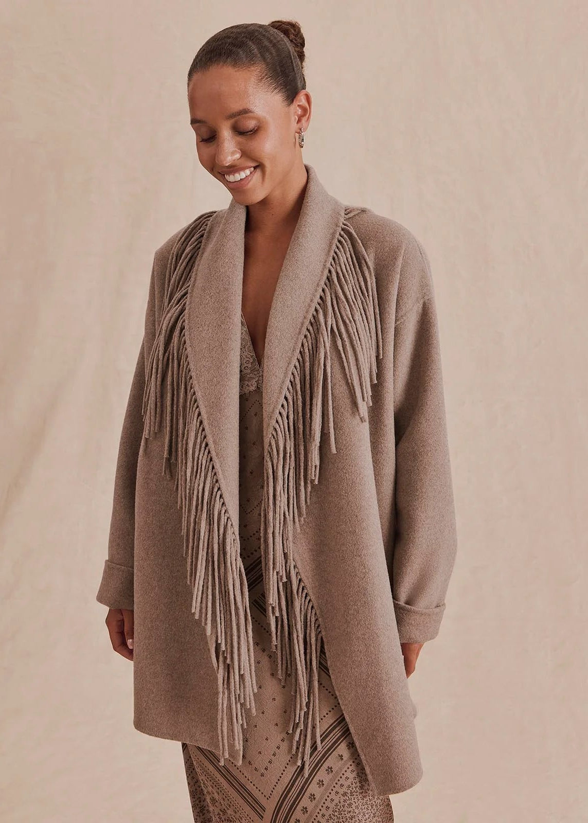 A woman wearing a taupe-brown fringed cardigan over a patterned dress, standing against a neutral beige backdrop.