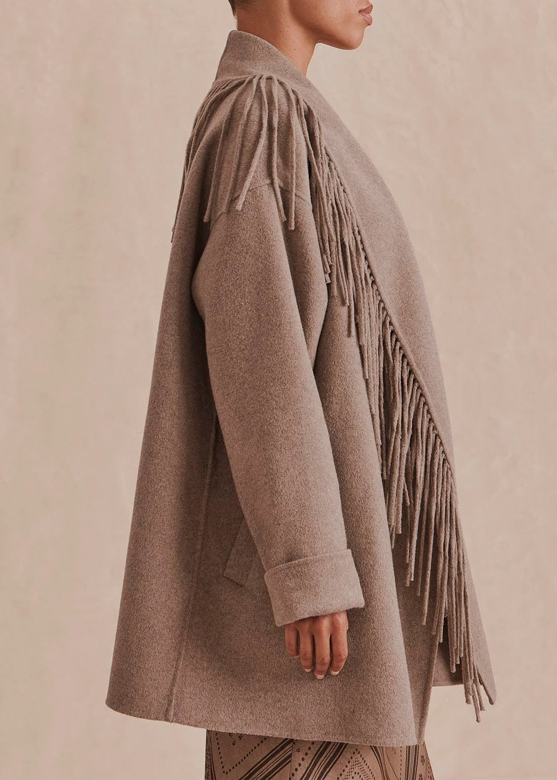A side-profile view of a model wearing a taupe-brown fringed wrap coat against a textured beige background.