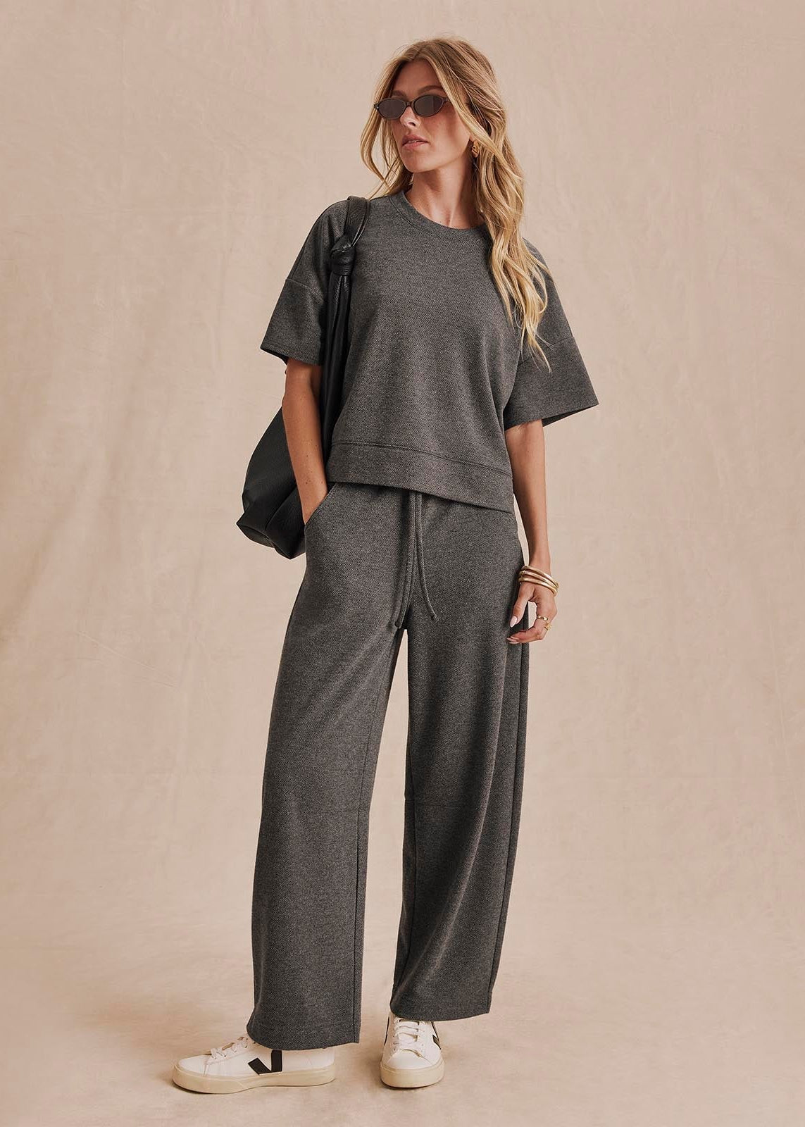 A woman models a dark gray two-piece lounge set consisting of a short-sleeve cropped t-shirt and matching wide-leg sweatpants, styled with white sneakers, sunglasses, and a black shoulder bag against a beige backdrop.