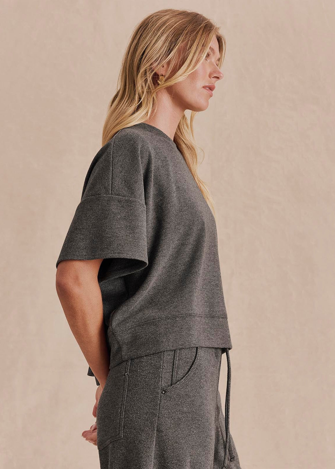 A woman in profile wearing a dark gray short-sleeve knit top and matching gray drawstring pants against a warm beige studio background.