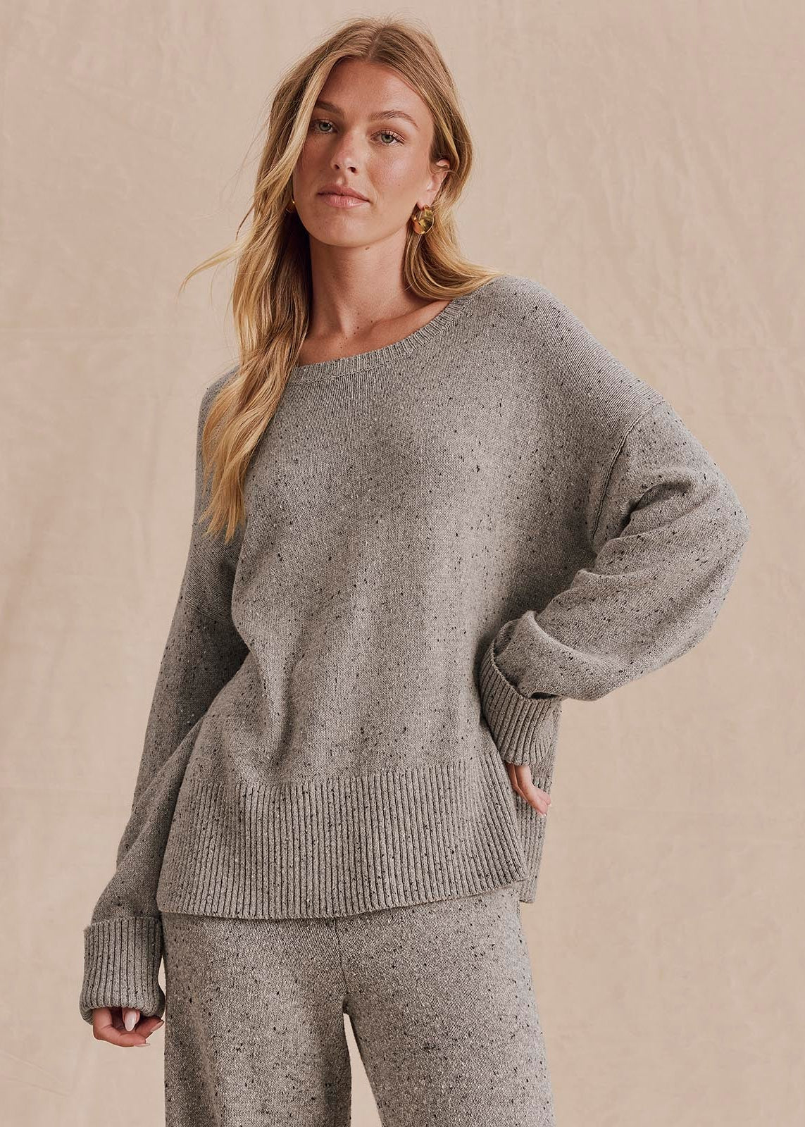 A woman modeling a light gray knit sweater and matching knit pants against a warm beige studio backdrop.