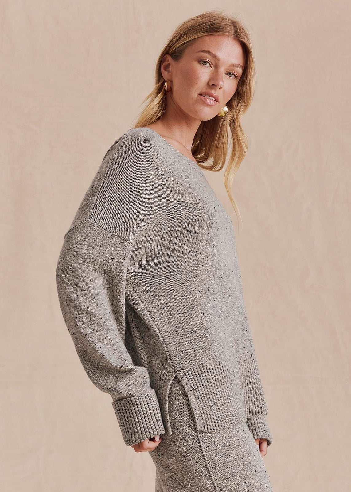 A woman wearing a light gray speckled knit sweater with a relaxed fit, ribbed cuffs, and a split hem, posed against a neutral beige textured background.