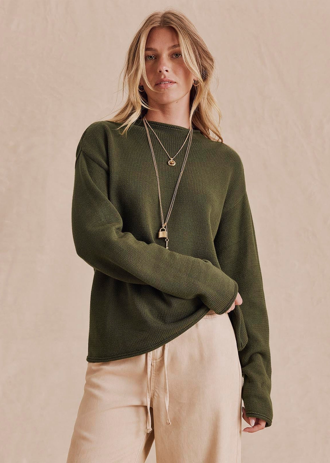 A woman wearing a dark olive green knit sweater and light beige drawstring pants, accessorized with gold necklaces, standing against a textured beige backdrop.