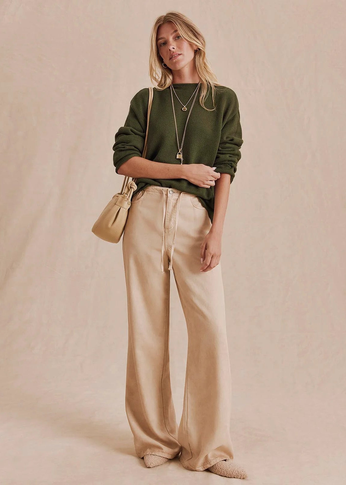 A woman stands against a warm beige backdrop wearing an olive green long-sleeve top, wide-leg beige pants, layered gold necklaces, and carrying a small tan shoulder bag.