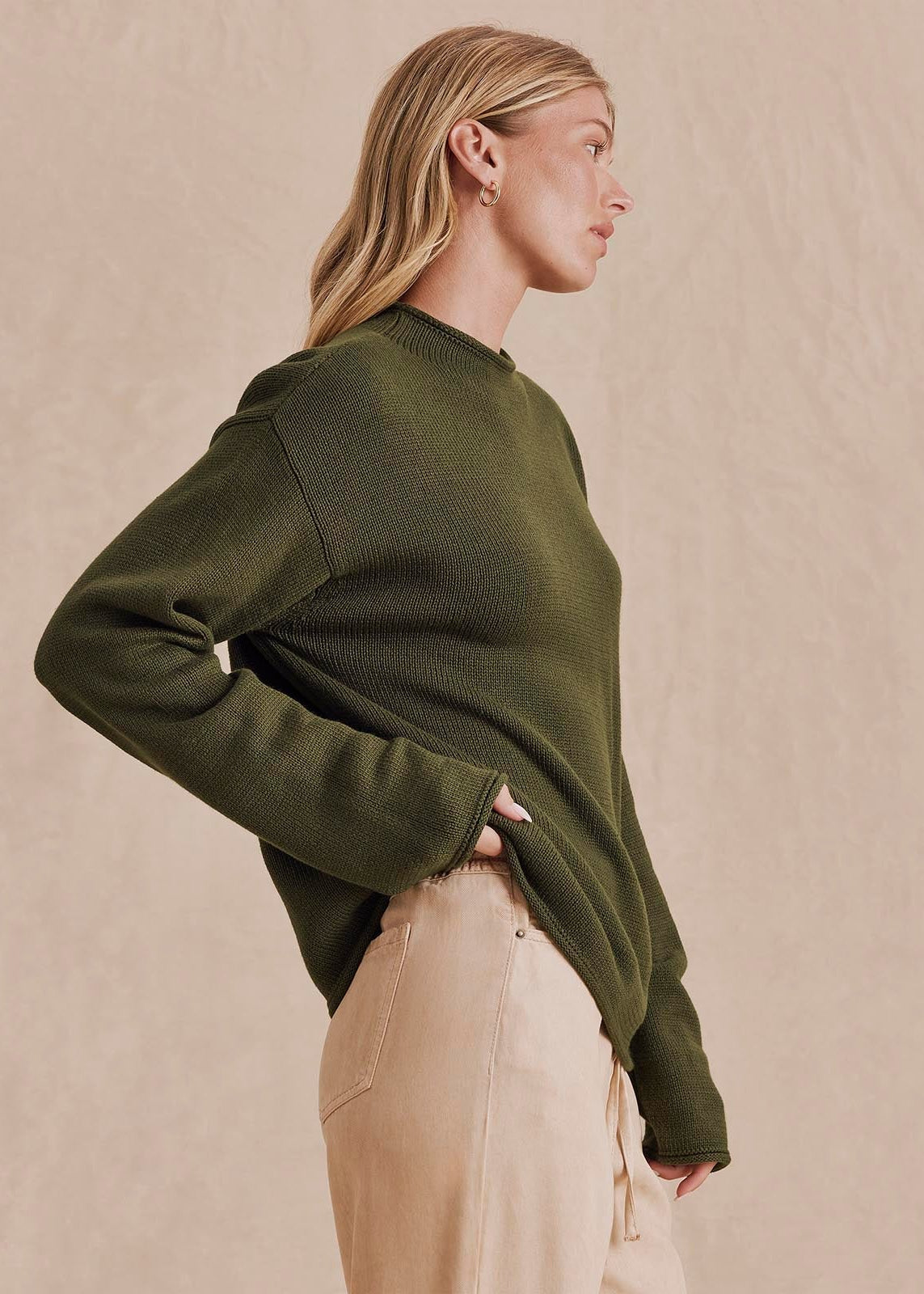 A woman in profile wearing an olive green ribbed knit sweater and beige trousers, standing against a textured beige studio background.