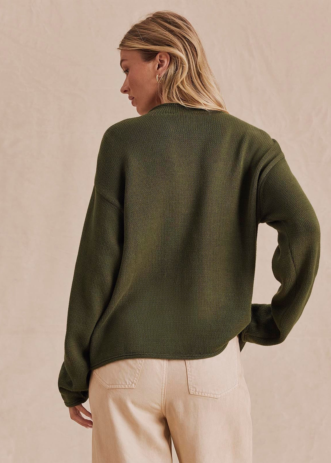 A woman shown from the back wearing an olive green knit sweater and light beige pants, standing against a textured beige wall.