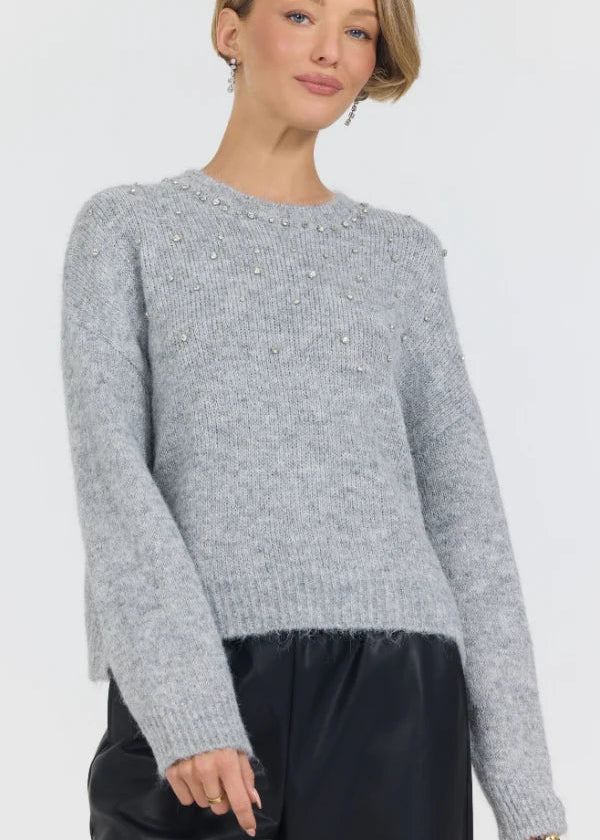 A woman wearing a light gray heathered knit sweater with delicate pearl embellishments across the chest, paired with black pants, against a plain light gray studio background.