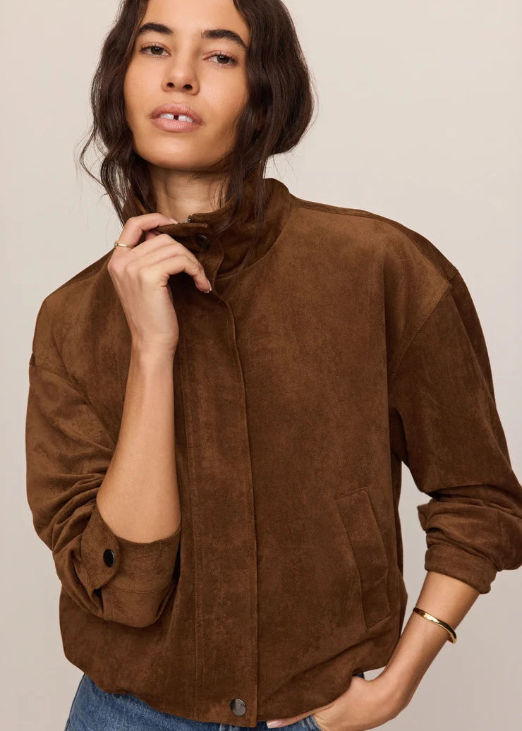 A woman wearing a brown suede jacket with a high collar and button-front closure, paired with blue jeans, photographed against a plain light beige studio background.
