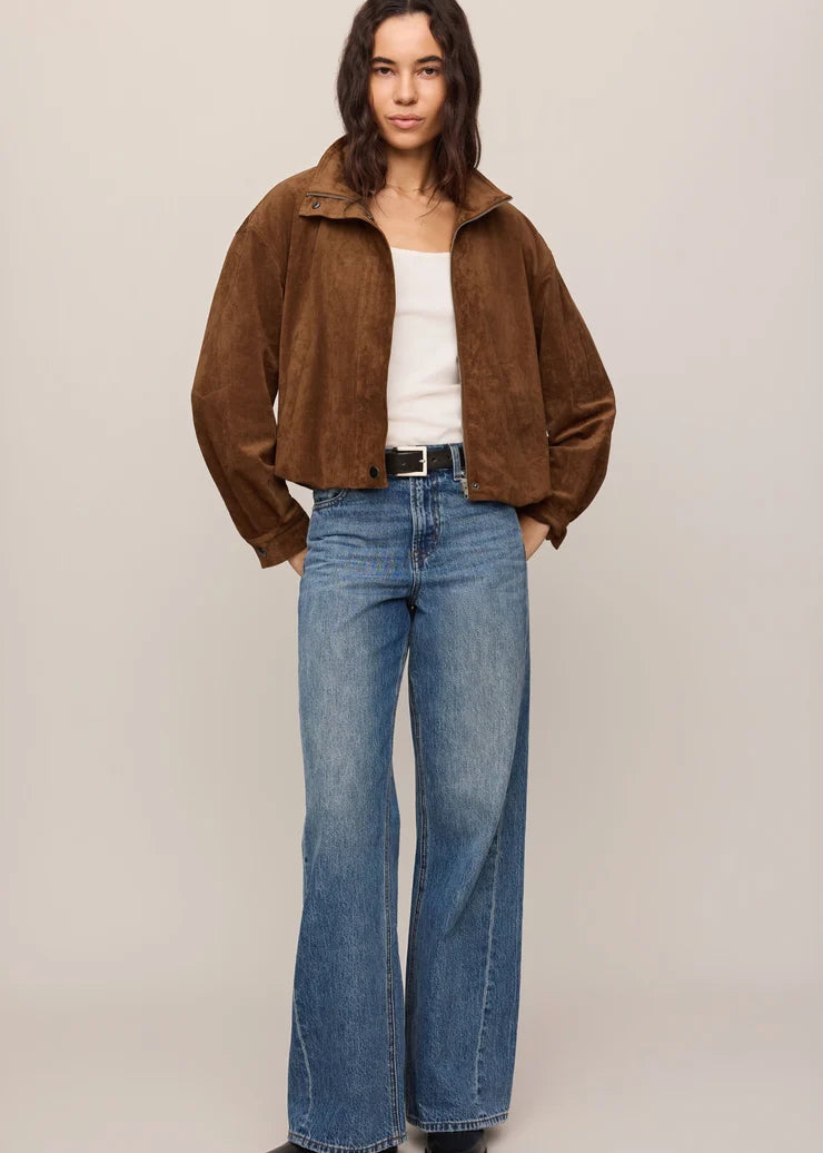 A woman modeling a brown suede jacket, blue wide-leg jeans, a white tank top, and black shoes against a plain beige studio background.