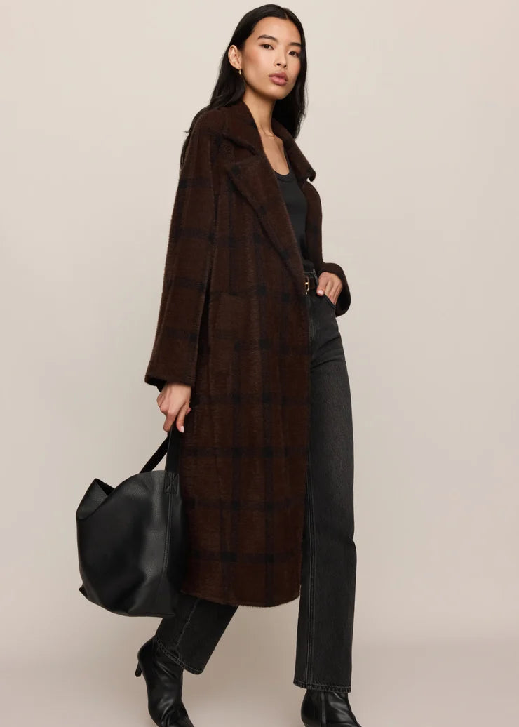 A model wearing a long brown plaid coat over a black outfit, holding a black leather bag against a plain beige studio background.