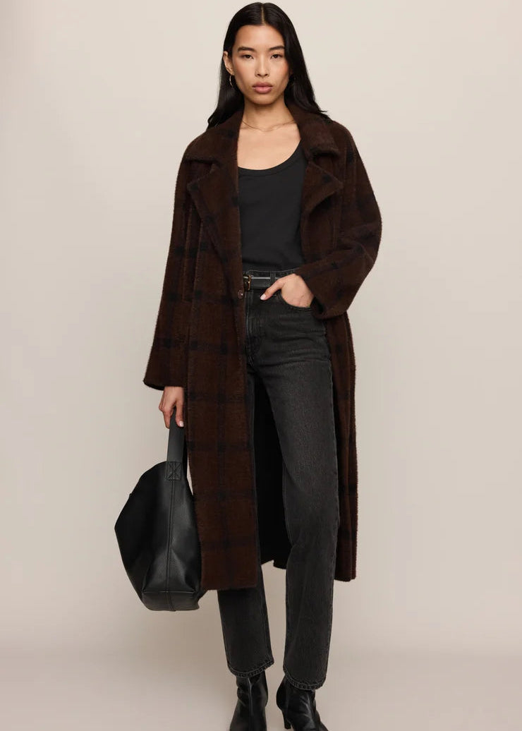 A model stands against a plain beige studio background wearing a long brown plaid coat over a black top and dark gray jeans, holding a black tote bag.