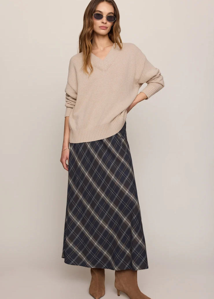 A woman modeling a beige knit V-neck sweater and a navy blue plaid maxi skirt, paired with brown ankle boots and sunglasses, against a plain light beige studio background.