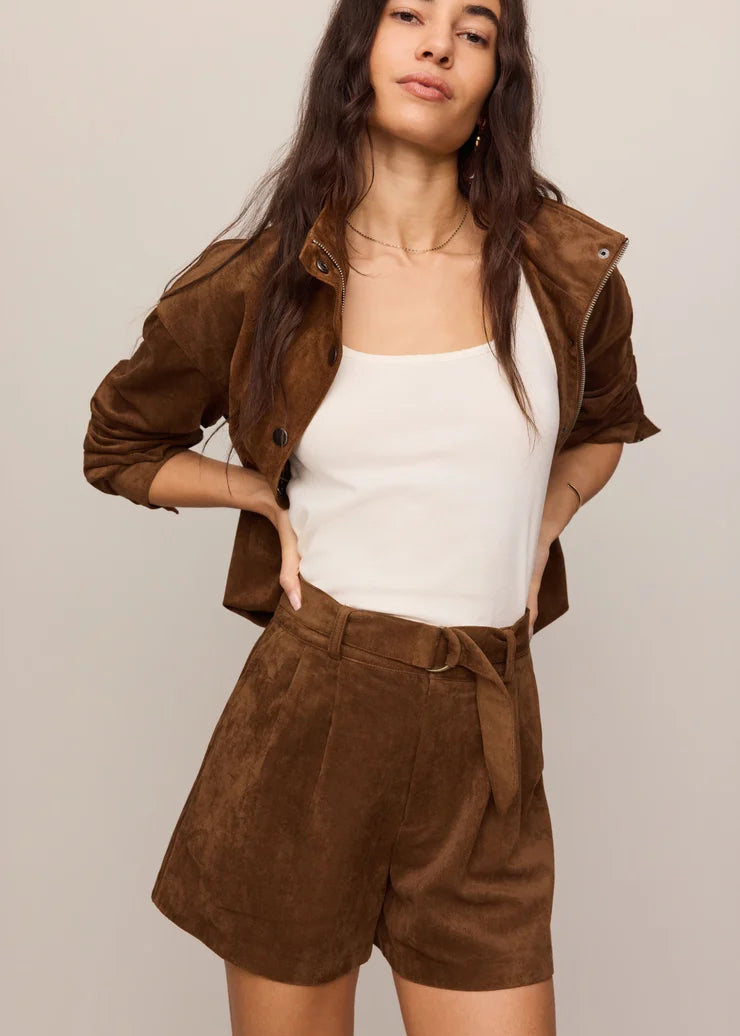 A woman modeling a brown suede jacket and matching brown suede belted shorts over a white tank top, photographed against a plain light gray studio background.
