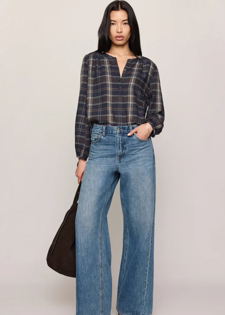 A model stands against a light gray studio background wearing a dark navy and black plaid long-sleeve blouse tucked into high-waisted, wide-leg blue jeans. She is holding a dark brown shoulder bag and wearing black pointed-toe flats.