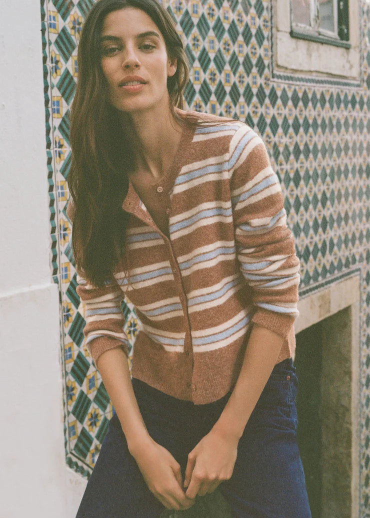 A woman wearing a brown, beige, and white striped knit cardigan and dark blue jeans, leaning against a wall with a green and white patterned tile design.