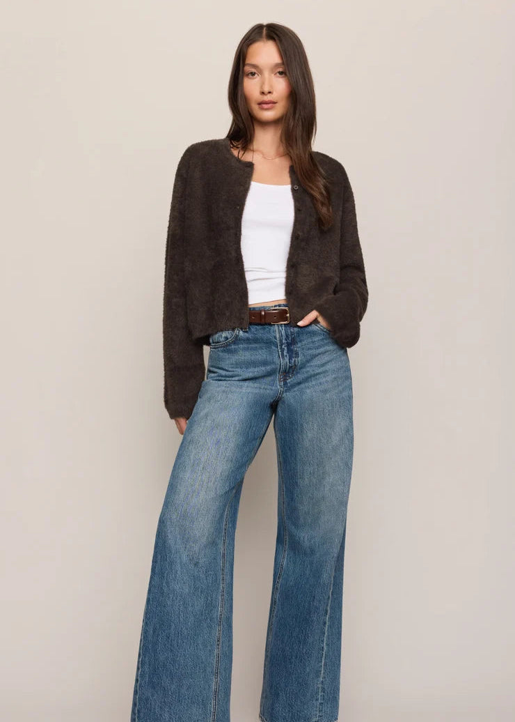 A woman modeling a dark brown fuzzy cropped cardigan, blue wide-leg jeans, a white tank top, and brown boots against a plain beige studio background.