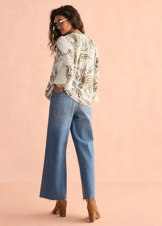 A woman wearing a white long-sleeve blouse with a brown and gold botanical pattern, paired with wide-leg blue jeans and tan ankle boots, photographed against a solid peach-pink studio background.