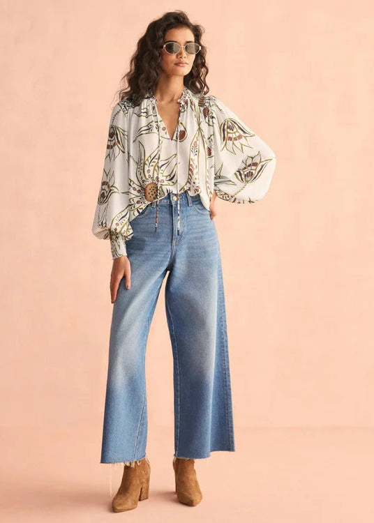 A woman modeling a white long-sleeve blouse with a brown and green floral pattern, paired with high-waisted wide-leg blue jeans and tan ankle boots, set against a solid peach-pink studio background.