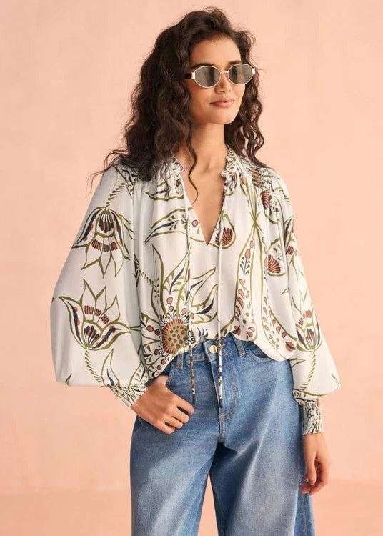 A woman wearing a white long-sleeve blouse with a brown and olive botanical print, styled with blue jeans and round sunglasses, against a solid peach-pink background.