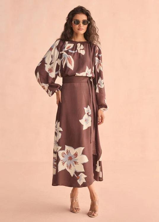 A woman modeling a brown midi dress with a white floral print, featuring a smocked bodice, long sleeves, and a matching tie belt, set against a solid peach-pink studio background.