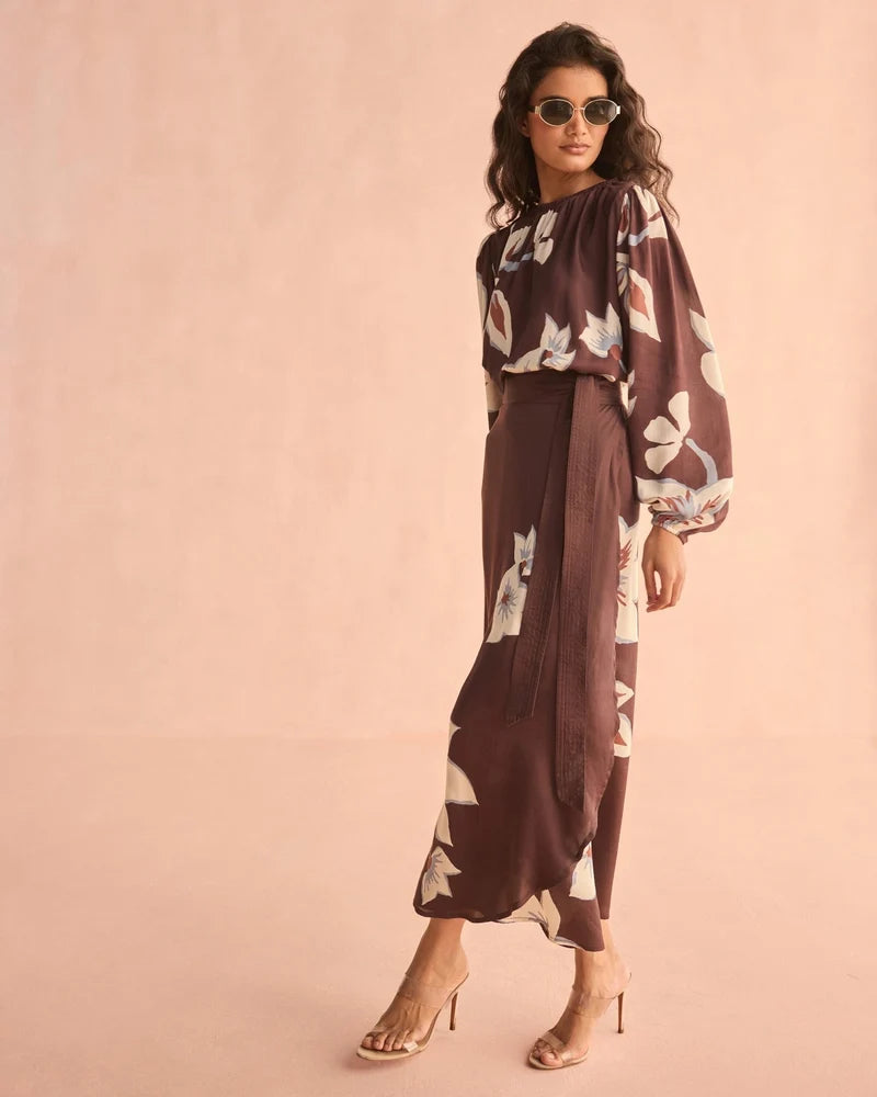 A woman modeling a brown two-piece set featuring a high-neck long-sleeve blouse and matching high-waisted pleated maxi skirt, both decorated with a white floral pattern. She is wearing beige high-heeled sandals and sunglasses against a solid light pink background.