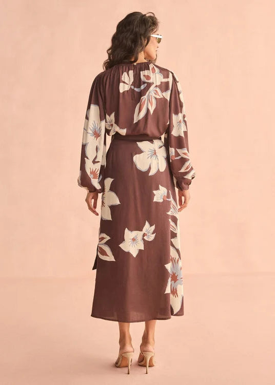 A woman viewed from the back wearing a long-sleeve brown maxi dress with a white floral print, standing against a solid light peach-pink background.