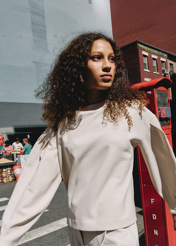 A woman with curly dark hair wearing a cream-colored long-sleeve top and matching pants, standing outdoors next to a red telephone booth with a city street and buildings in the background.