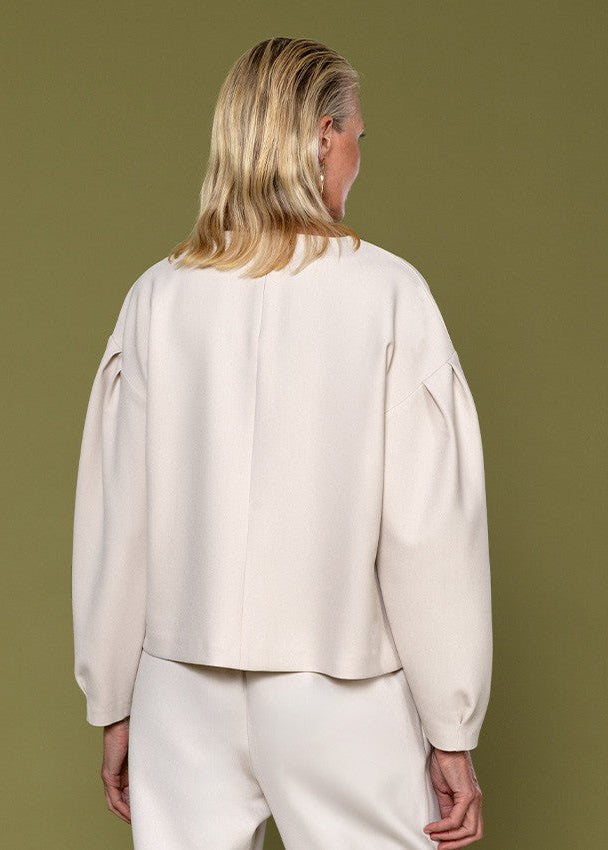 A woman shown from the back wearing a cream-colored cropped long-sleeve blouse with dramatic puffed sleeves and matching cream trousers, set against a solid olive green background.