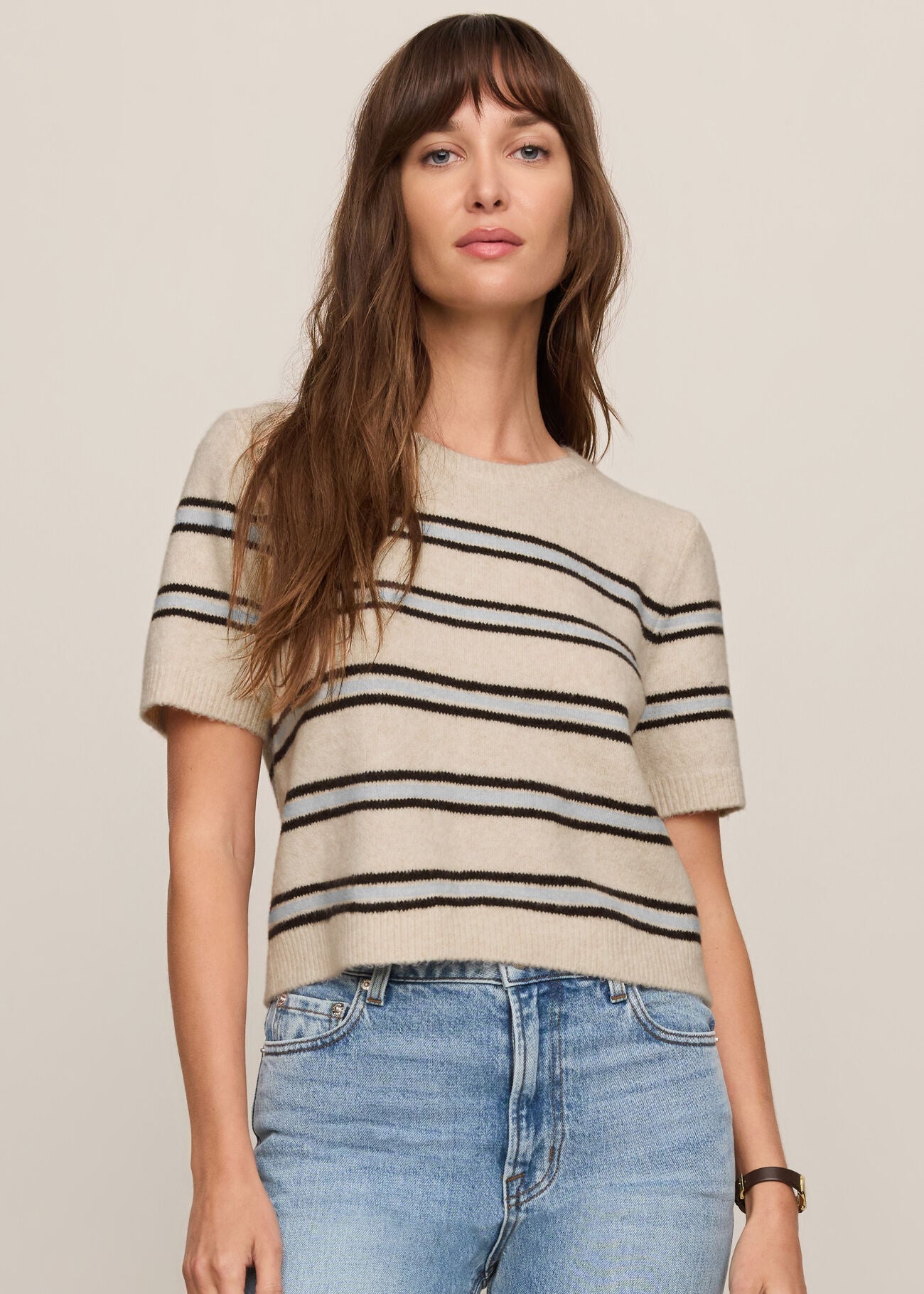 Woman wearing a beige sweater with black stripes and blue jeans on a plain background