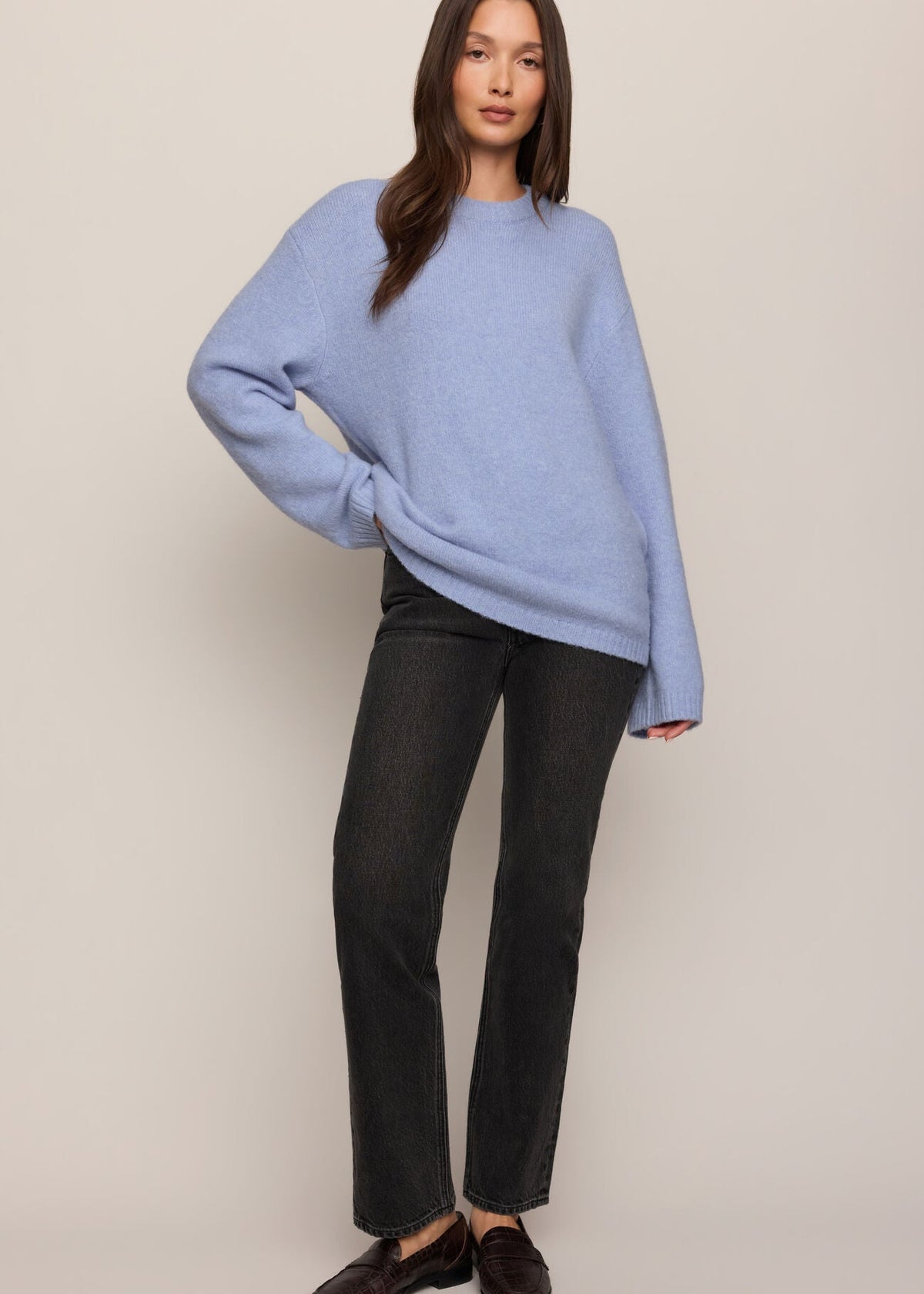 A woman modeling an oversized light blue knit sweater, paired with dark charcoal jeans and dark brown leather loafers, against a solid light beige studio background.