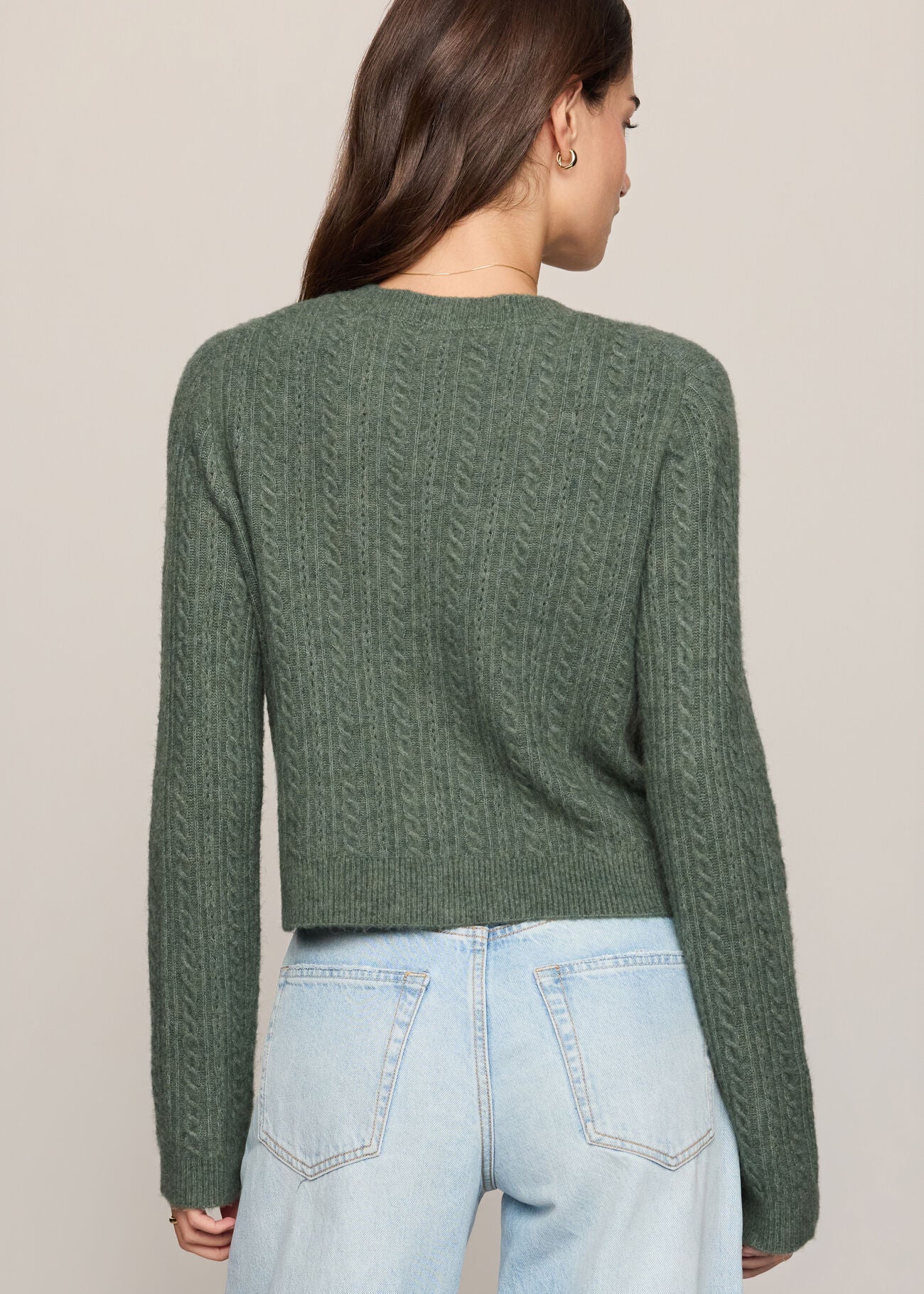 A woman viewed from the back wearing a sage green cable-knit cropped sweater and light-wash blue jeans against a plain beige studio background.