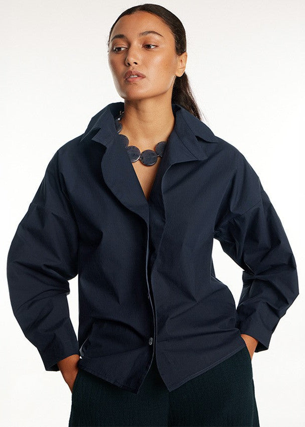 A woman modeling a dark navy blue oversized button-up shirt with a relaxed fit, paired with dark pants and a chunky dark necklace, against a plain white studio background.