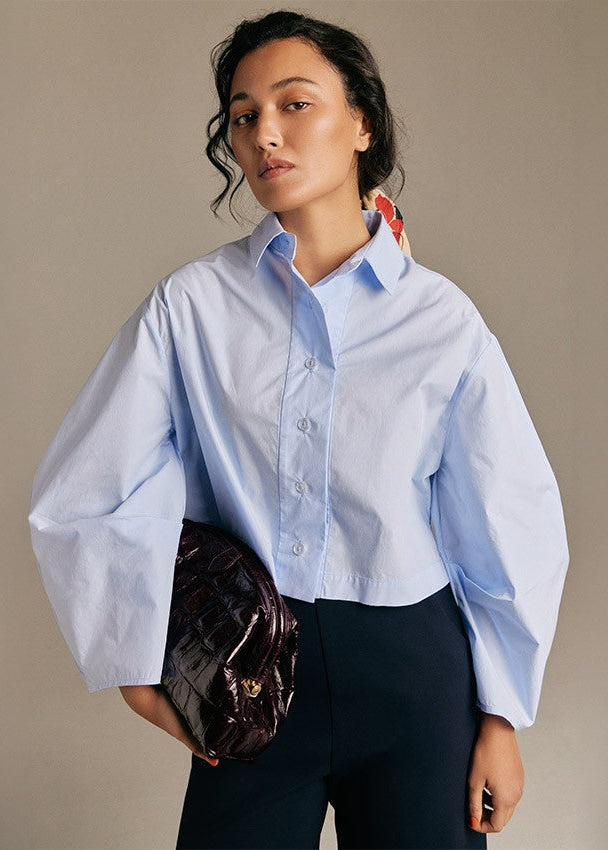 A model wearing a light blue cropped button-down shirt with dramatic balloon sleeves, paired with dark trousers and holding a dark metallic clutch bag against a neutral beige background.