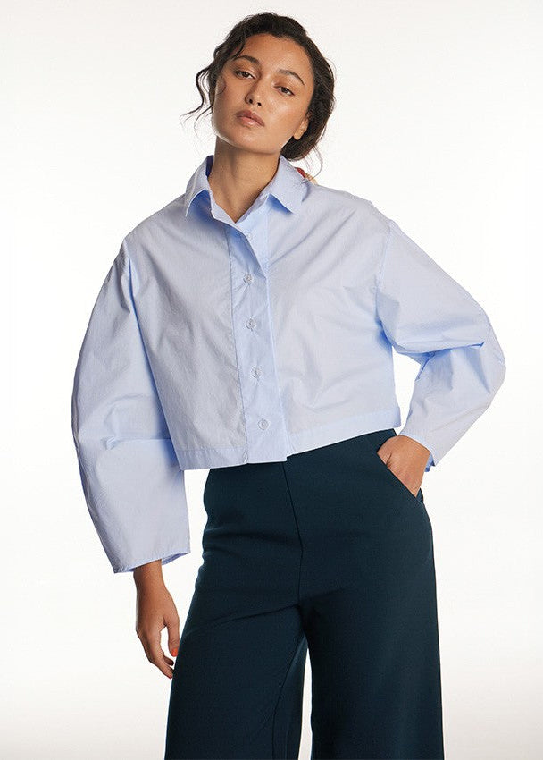 A woman modeling a light blue cropped button-up shirt with voluminous long sleeves, paired with dark teal trousers, against a plain white studio background.
