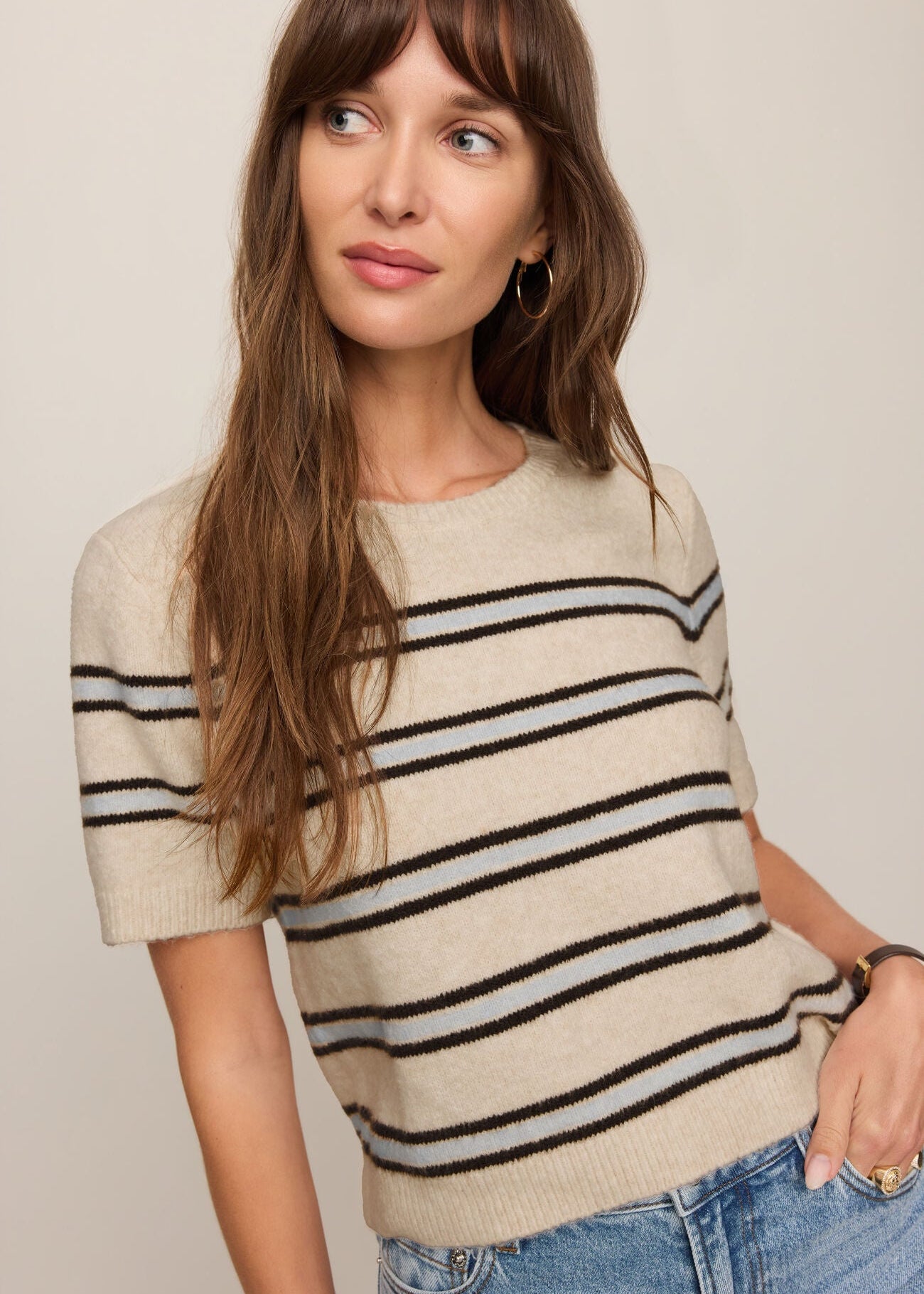 Woman wearing a beige and black striped sweater with blue jeans against a plain background