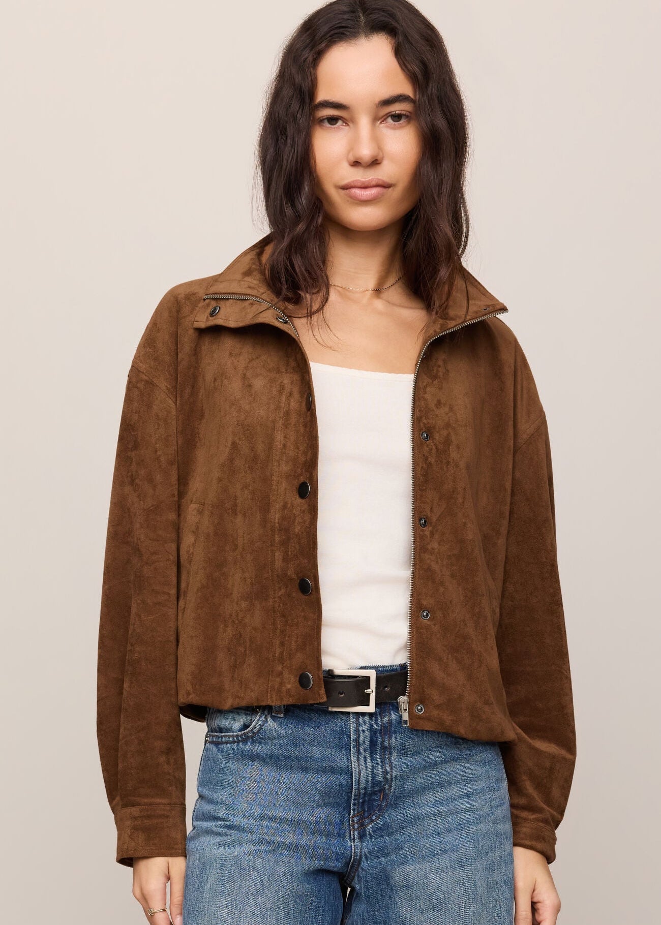 Woman wearing a brown suede jacket over a white top and blue jeans on a beige background