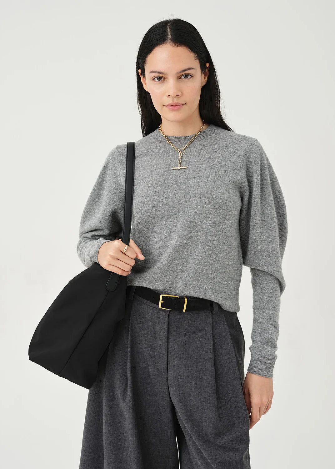 A woman modeling a heather gray long-sleeve knit top with draped sleeves, paired with dark gray wide-leg trousers, a black belt with a gold buckle, and carrying a black shoulder bag against a light gray studio background.