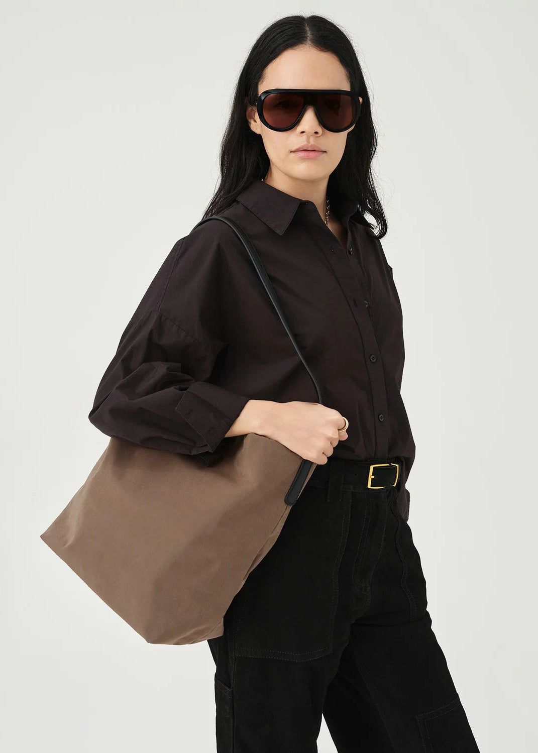 A model wearing oversized dark sunglasses, a dark brown button-up shirt, and black pants carries a large taupe-brown canvas tote bag over her shoulder against a light gray studio background.