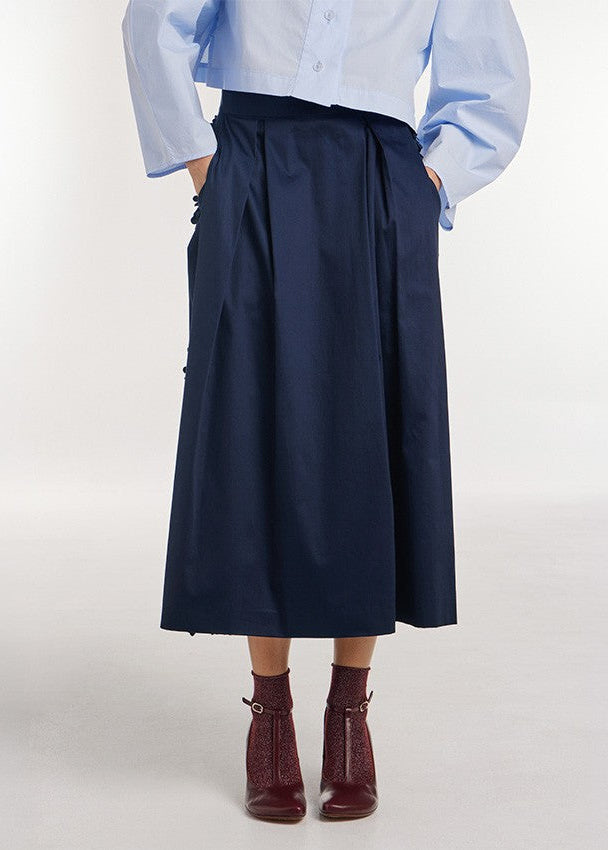 A model is shown from the waist down wearing a dark navy blue high-waisted midi skirt with side pockets, paired with a light blue button-down shirt and dark red ankle boots.
