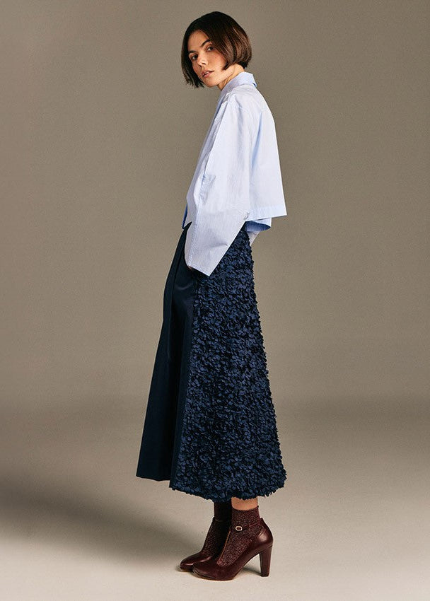 A model is shown from the side wearing a light blue button-down shirt, a dark navy textured midi skirt, and dark brown heeled ankle boots, set against a solid warm gray studio background.