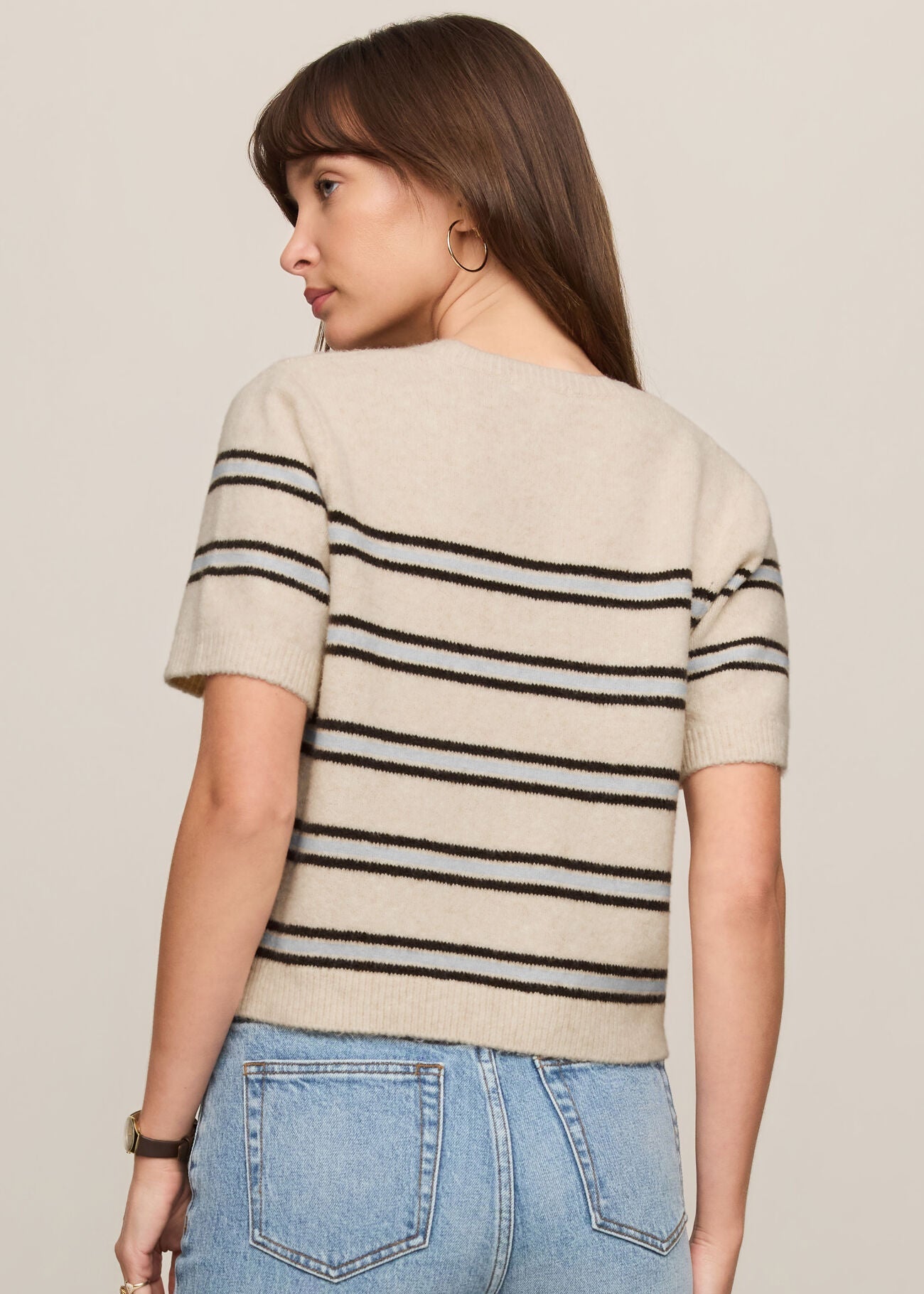 Woman wearing a beige sweater with black stripes and blue jeans on a plain background